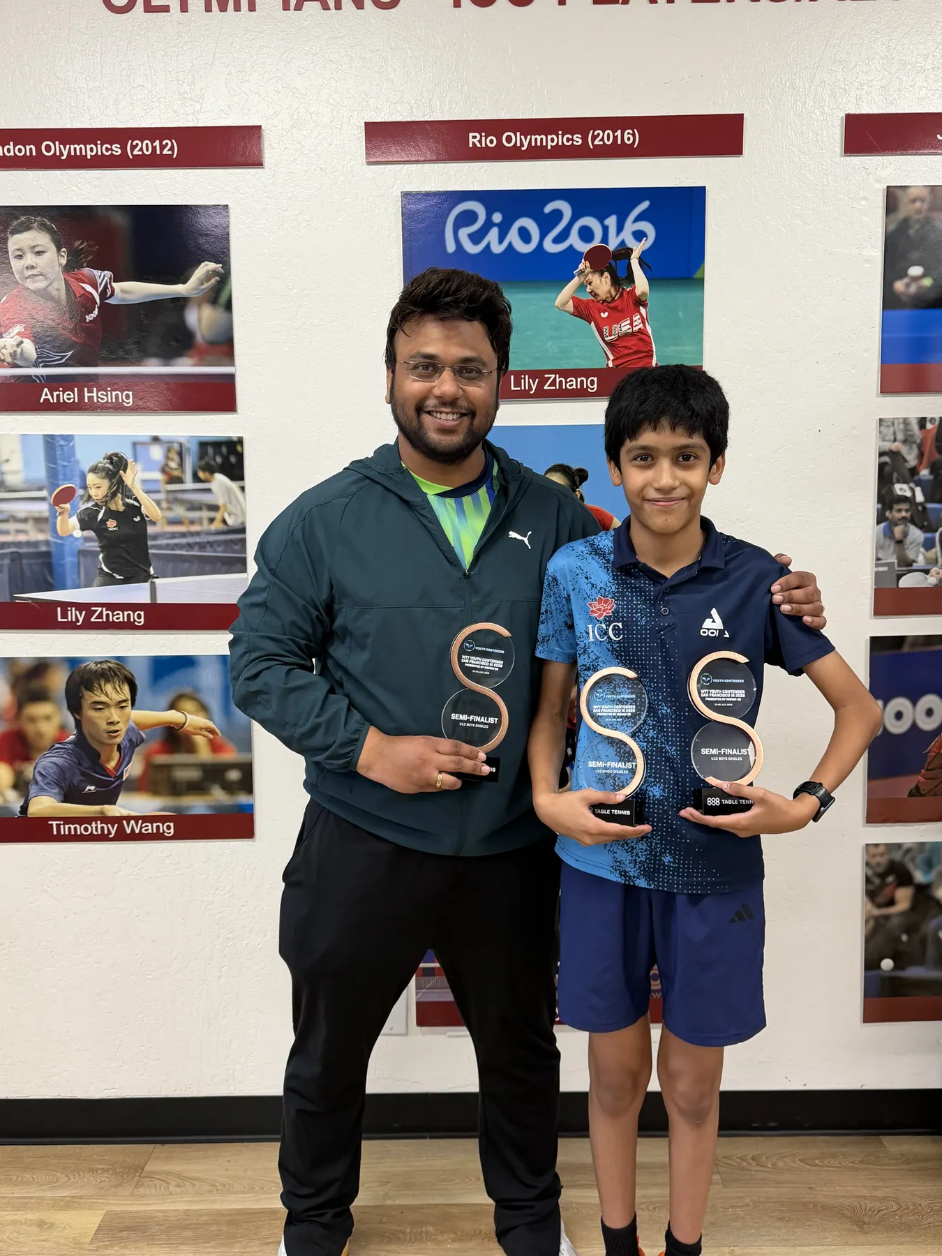Shreyan Modak and Subham Kundu holding semifinalist trophies in front of the ICC Olympians wall