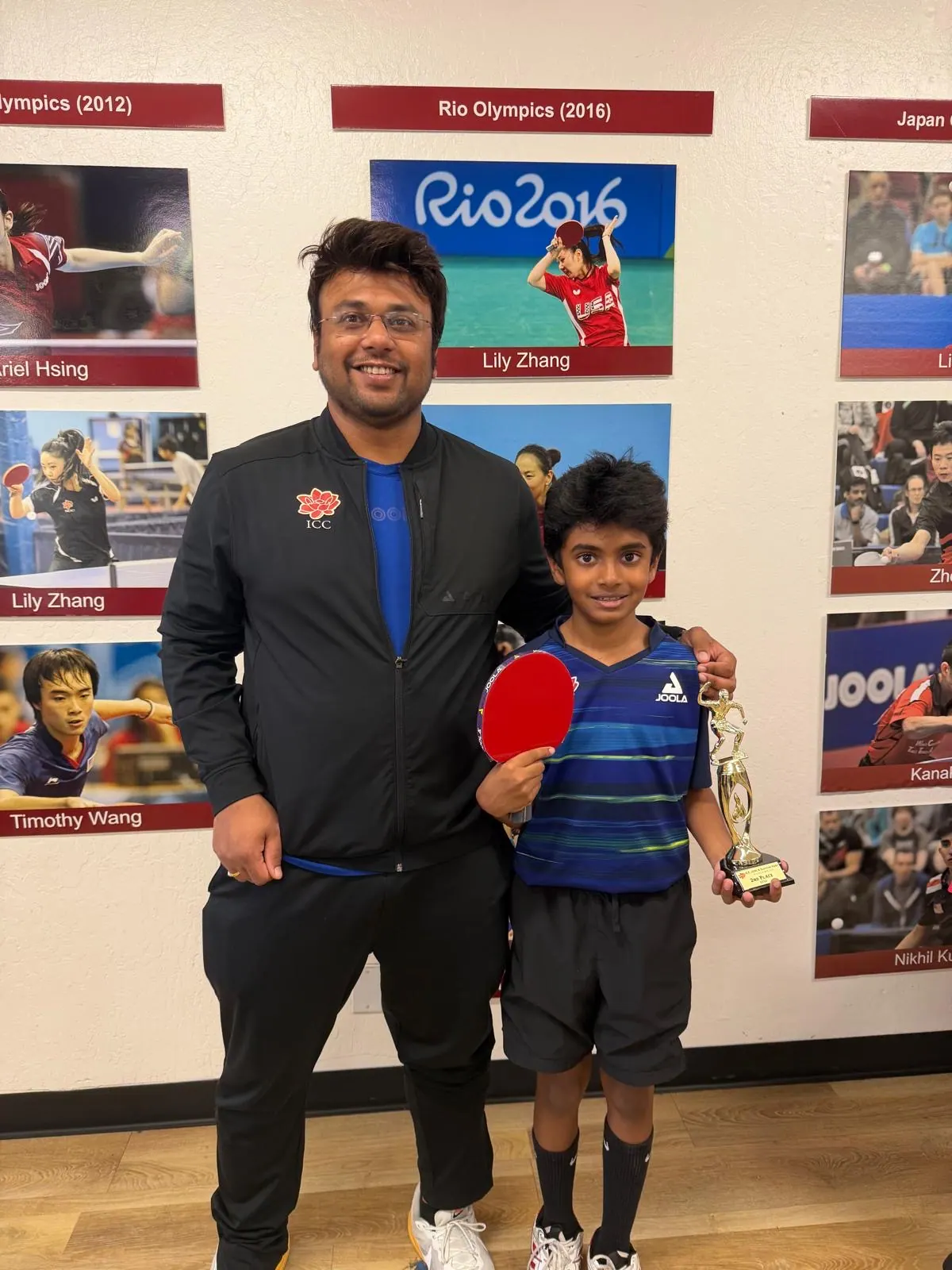 Adhwin Sasikumar holding his bat and trophy beside Subham Kundu in front of the ICC Olympians wall
