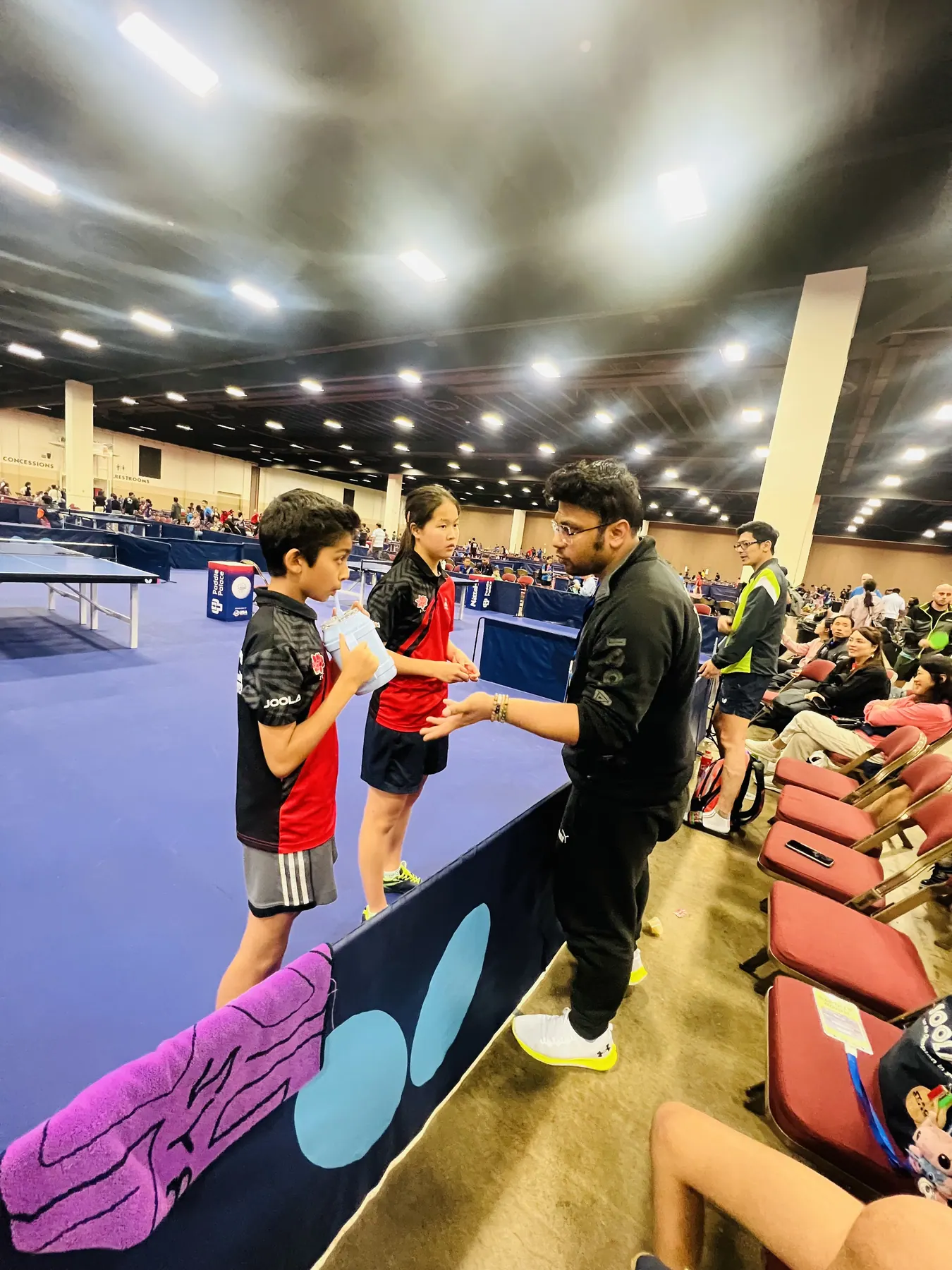 Subham Kundu coaching two young players courtside at a large tournament venue