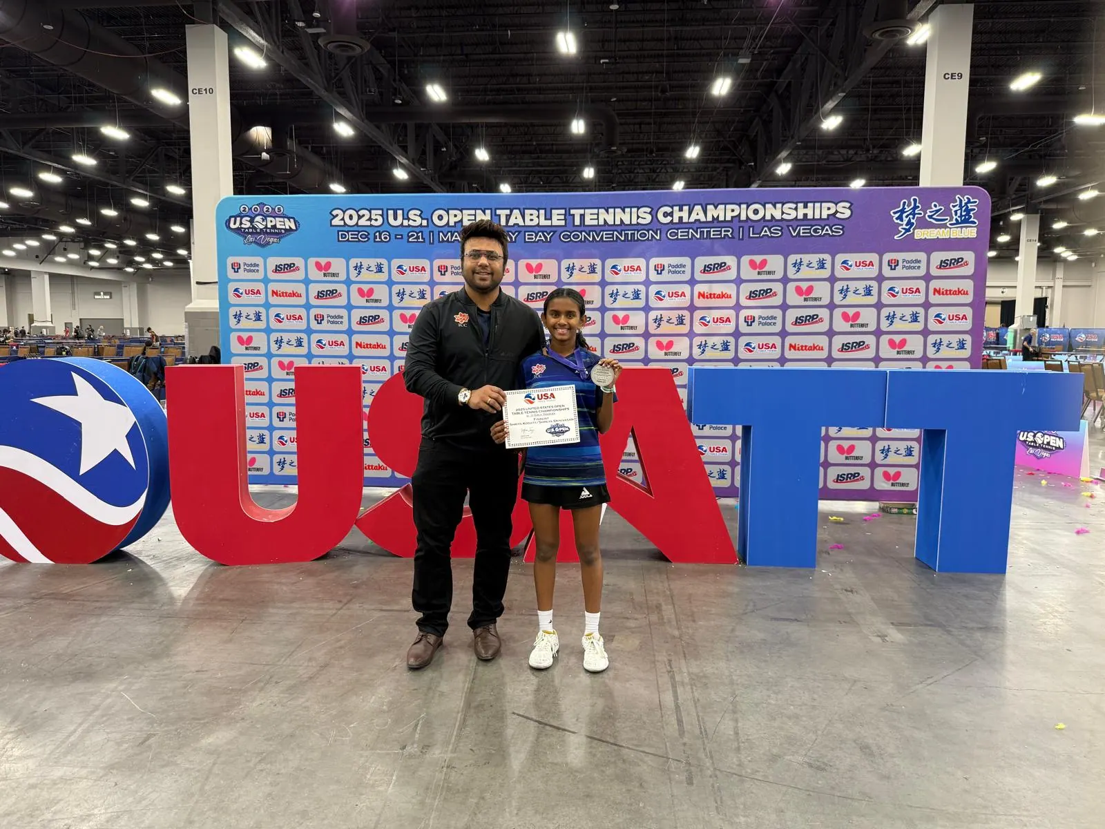 Shriya Kodaty with her silver medal and US Open certificate beside Subham Kundu in front of the USATT letters at the 2025 US Open